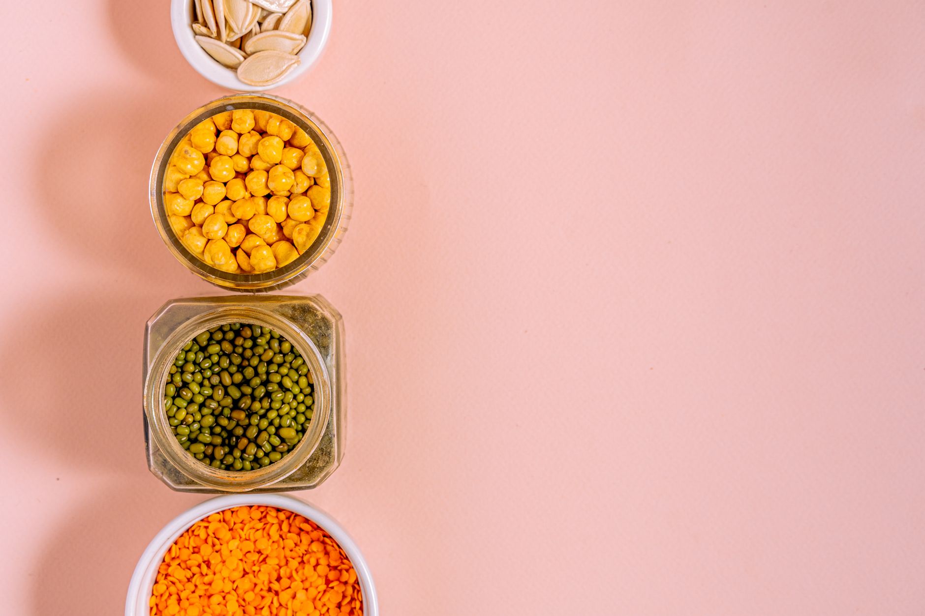 Flat lay of chickpeas, lentils, mung beans, and pumpkin seeds with pink backdrop.
